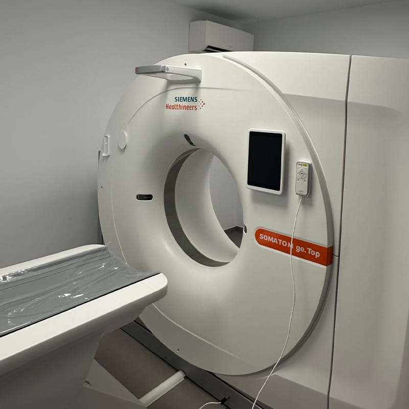 Un scaner CT Siemens Healthineers SOMATOM go.Top într-o cameră a Clinicii Medicala Grama, cu o masă de pacient acoperită cu plastic poziționată în fața deschiderii circulare a scanerului.