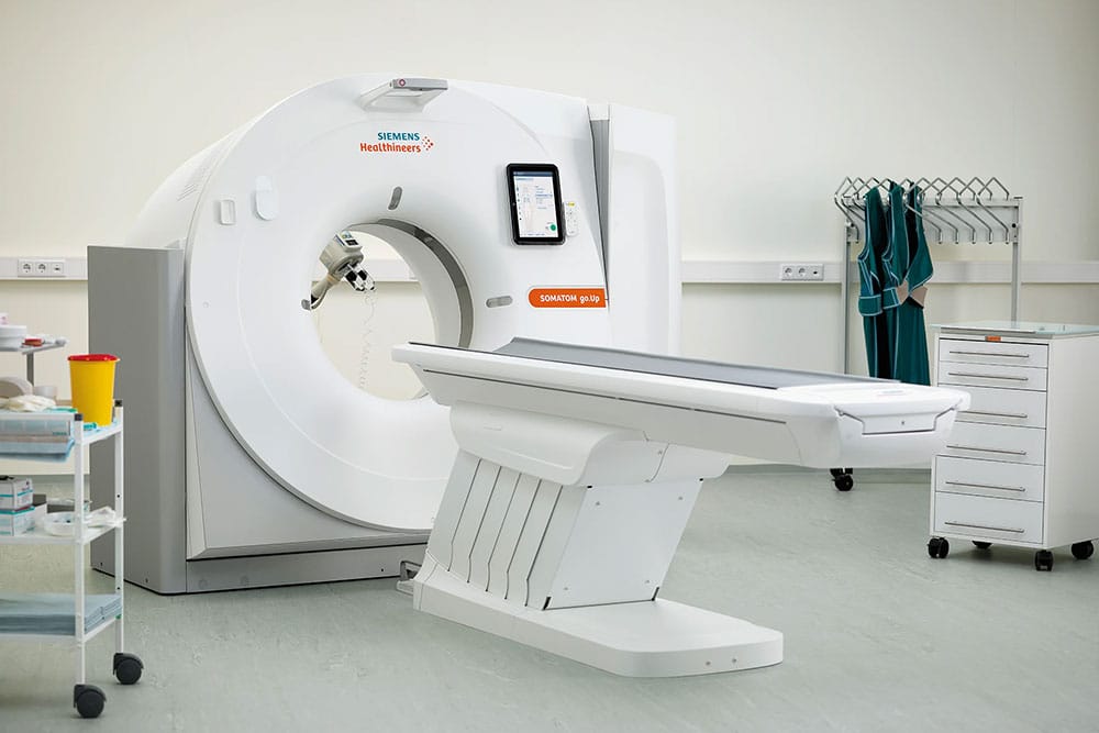 Un scaner CT modern cu o masă pentru pacient într-o cameră medicală curată și luminoasă la Clinica Medicala Grama. Echipamentul medical, cărucioarele de depozitare și șorțurile de plumb sunt vizibile în fundal.
